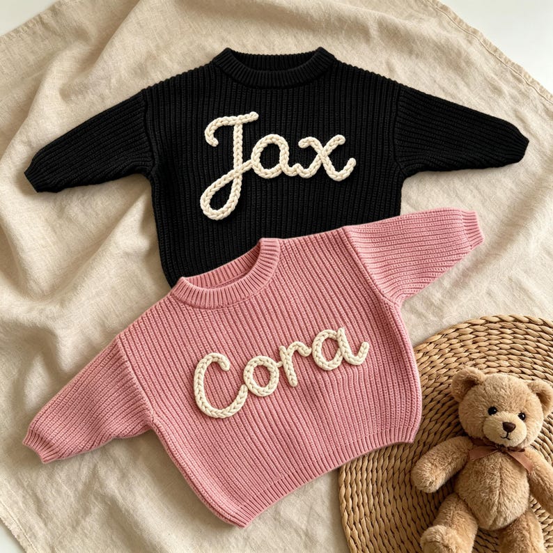 May include: A black long-sleeve knit sweater with the name Jax in white rope lettering and a pink long-sleeve knit sweater with the name Cora in white rope lettering. Both sweaters feature a ribbed crew neckline and a thick, textured knit pattern.