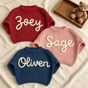 May include: Three long-sleeve knit sweaters in red, pink, and blue. Each sweater features a crew neckline and a ribbed texture. The front of each sweater displays a name in raised, white, braided yarn lettering: Zoey, Sage, and Oliven.