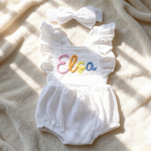 Puede incluir: Romper de beb&eacute; blanco con mangas con volantes y diadema a juego. El romper presenta el nombre "Elsa" bordado con hilo colorido. El conjunto est&aacute; hecho de una tela suave y texturizada.