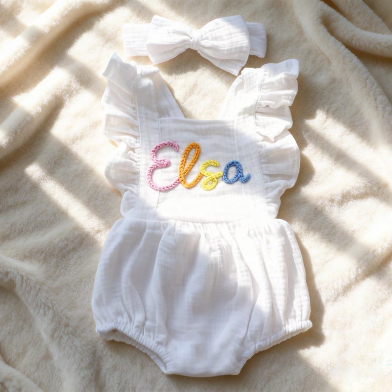Puede incluir: Romper de beb&eacute; blanco con mangas con volantes y diadema a juego. El romper presenta el nombre "Elsa" bordado con hilo colorido. El conjunto est&aacute; hecho de una tela suave y texturizada.