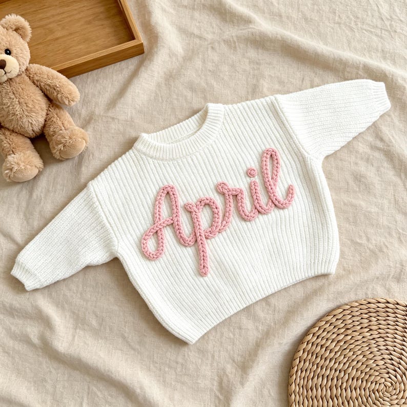 May include: A white long-sleeved ribbed sweater featuring the name April embroidered in pink yarn across the chest. The sweater has a crew neckline and a textured knit pattern, presented alongside a brown teddy bear and a woven circular mat.