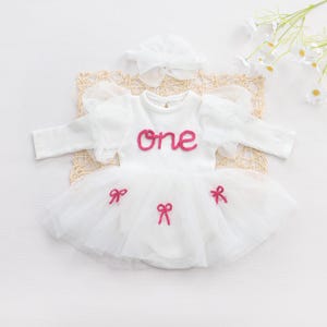 May include: White baby outfit with a tutu skirt and long sleeves. The word "ONE" is embroidered in pink on the chest. The outfit includes a matching headband and pink bow accents on the skirt. A perfect outfit for a baby's first birthday.