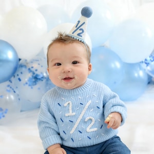May include: A baby wearing a light blue sweater with the number "1/2" in white and a matching party hat. The background features blue and white balloons, creating a festive atmosphere for a half-birthday celebration.