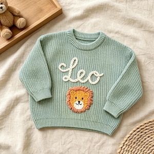 May include: A light green long-sleeve knit sweater featuring a white embroidered script text that reads Leo, positioned above a circular, textured applique of a lion face with a yellow center and a light brown mane.