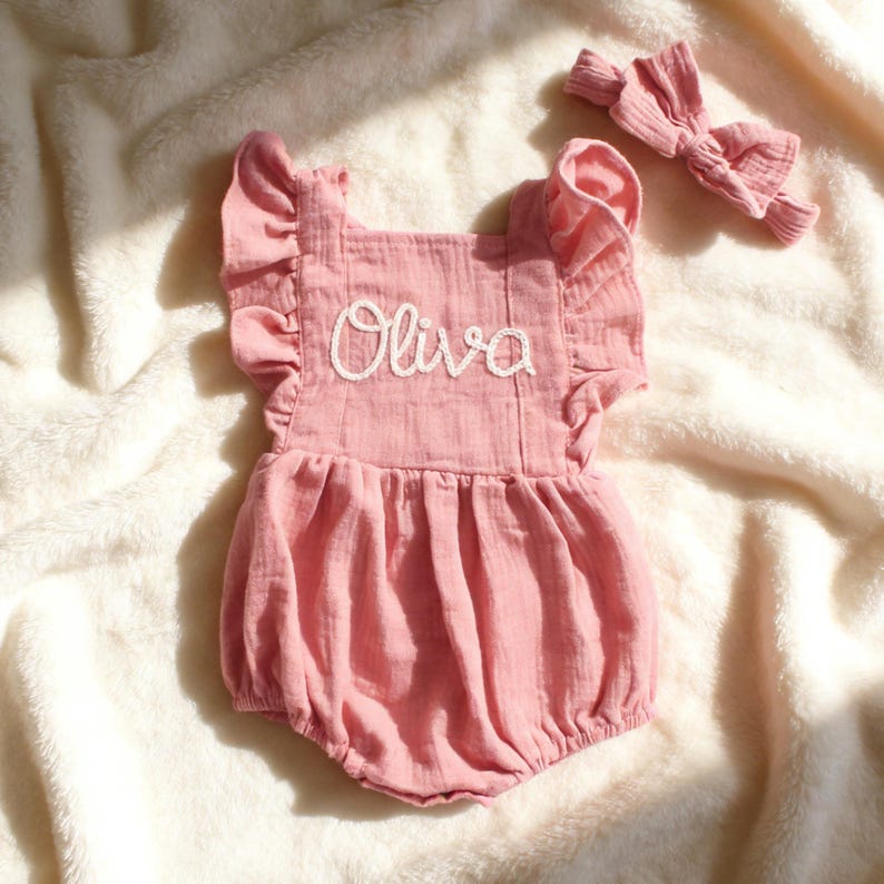 Puede incluir: Un pelele rosa para beb&eacute; con el nombre "Olivia" bordado en escritura blanca. El pelele tiene mangas con volantes y una diadema a juego. La tela parece ser un material suave y texturizado.