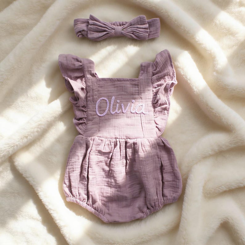 Puede incluir: Un conjunto de ranita y diadema para beb&eacute; de color malva. La ranita presenta mangas con volantes y el nombre "Olivia" bordado en la parte delantera. El conjunto est&aacute; hecho de una tela suave y texturizada y se muestra sobre una manta blanca y esponjosa.