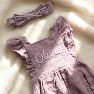 Puede incluir: Conjunto de beb&eacute; en color rosa empolvado, compuesto por un pelele y una diadema a juego. El pelele presenta mangas con volantes y el nombre "Olivia" bordado en una delicada escritura. El conjunto es de tejido suave y texturizado.