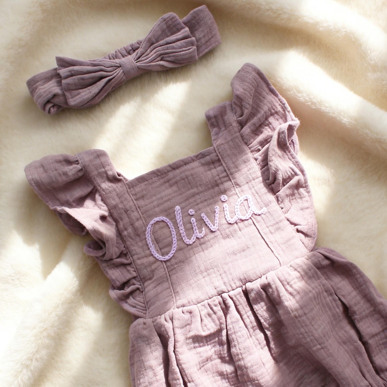 Puede incluir: Conjunto de beb&eacute; en color rosa empolvado, compuesto por un pelele y una diadema a juego. El pelele presenta mangas con volantes y el nombre "Olivia" bordado en una delicada escritura. El conjunto es de tejido suave y texturizado.