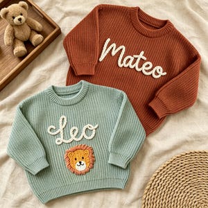 May include: A rust-colored knit sweater with the name Mateo embroidered in white script and a sage green knit sweater featuring the name Leo in white script above a small, embroidered lion face with a textured orange mane.