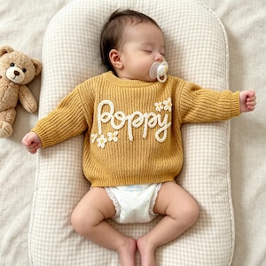 May include: A mustard yellow long-sleeve knit sweater featuring the white embroidered text Poppy and small white flower appliqu&eacute;s on the front. The sweater has a ribbed crew neckline, cuffs, and hem, providing a textured appearance for a baby.