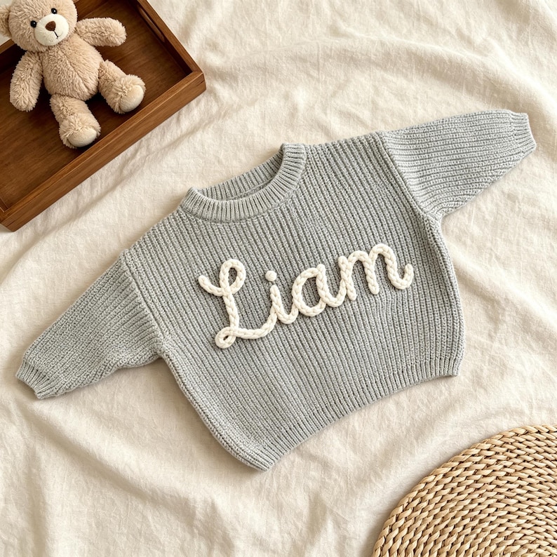May include: A light gray long-sleeve sweater featuring a ribbed knit texture and a crew neckline. The front of the sweater displays the name Liam in large, white, raised, braided yarn lettering stitched onto the center of the garment.