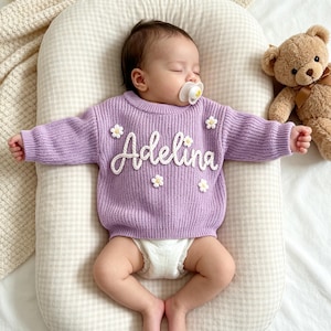 May include: A long-sleeved, ribbed knit sweater in a light purple color. The sweater features the name Adelina embroidered in white script across the chest, accented with small, white, three-dimensional daisy flower appliqu&eacute;s scattered around.