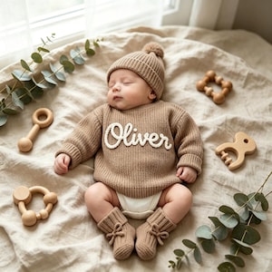 May include: A beige knitted long-sleeve sweater with the name Oliver embroidered in white script on the chest, paired with a matching beanie, ribbed booties with small bows, and a white diaper cover, surrounded by wooden baby rattles.