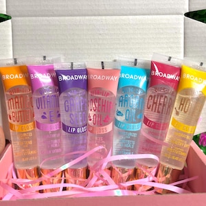 May include: A set of six lip gloss tubes in a pink box. Each tube has a different color cap and label, with flavors like Mango Butter, Vitamin E, and Cherry. The tubes are clear, showing the gloss inside.