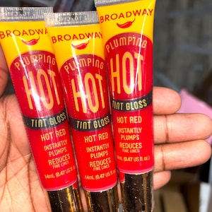 May include: Three tubes of Broadway Plumping Hot Tint Gloss in Hot Red. The tubes are yellow and red with gold caps. The text on the tubes reads "Instantly Plumps, Reduces Fine Lines" and "14ml (0.47 US fl. oz.)".