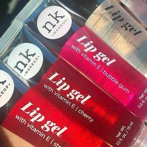 May include: Close-up of several tubes of lip gel in various colors. The tubes are labeled "Lip gel with vitamin E" and include flavors like cherry and bubble gum. The brand name "nk makeup" is visible on some tubes. Each tube contains 0.5 fl. oz. / 15 ml.
