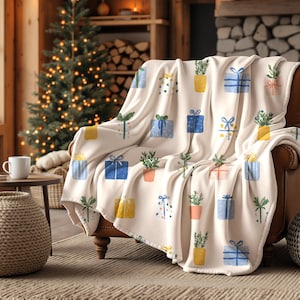 May include: A cream-colored throw blanket draped over a brown leather armchair. The blanket features a repeating pattern of colorful gift boxes and potted plants. A Christmas tree and fireplace are visible in the background.