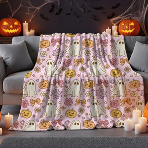 May include: A light pink throw blanket featuring a repeating pattern of white ghosts, orange jack-o'-lanterns, purple and white flowers, and tan bows. The blanket is draped over a gray sofa, showcasing a festive Halloween-themed design.