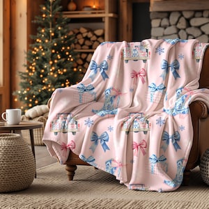 May include: A pink fleece blanket with a repeating pattern of gift boxes, trucks, bows, and snowflakes in blue, pink, and white. The blanket is draped over a brown armchair. A Christmas tree and a fireplace are in the background.
