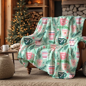May include: A soft throw blanket with a pale green backdrop, patterned with pink, white, and teal coffee mugs and green sprigs. The blanket is draped over a brown leather armchair in a living room setting, near a fireplace.