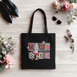 May include: Black tote bag with the text "MAMA needs WINE" in colorful block letters. The design includes a wine glass graphic. The bag is made of a sturdy fabric and has two black handles.