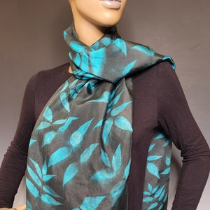 A black and teal silk scarf is arranged on the neck of a mannequin. The scarf has a black background with teals leaves.
