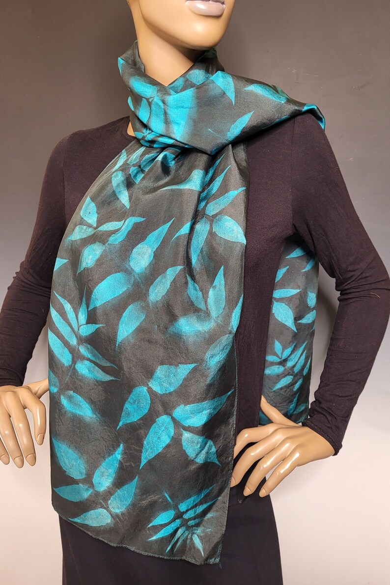 A black and teal silk scarf is arranged on the neck of a mannequin. The scarf has a black background with teals leaves.