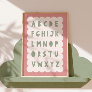 Pode incluir: Uma impressão alfabética emoldurada com fundo rosa e borda branca ondulada. As letras são na cor verde sálvia. A impressão é exibida em uma prateleira verde em forma de nuvem.