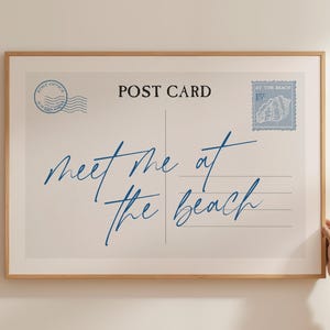 May include: A framed print resembling a vintage postcard. The design features the words "meet me at the beach" in blue script, along with postage stamps and the words "Post Card". The frame is a light brown color.