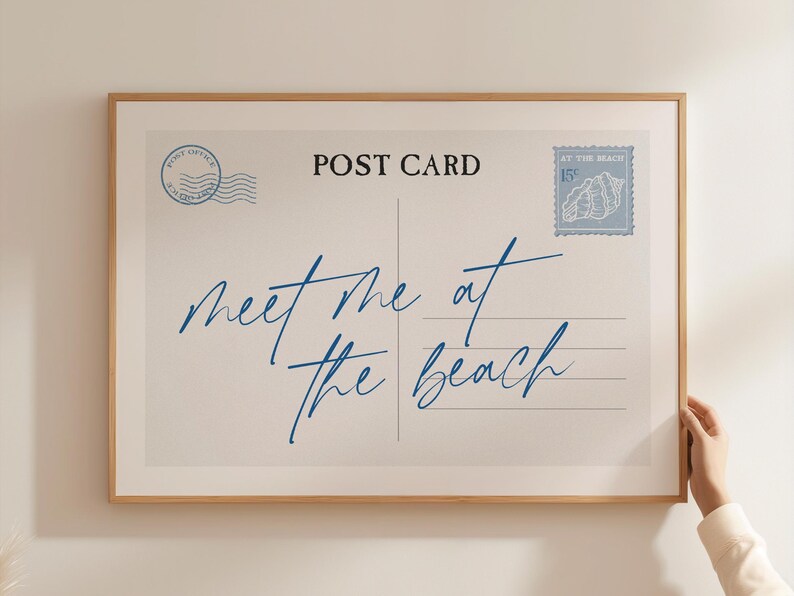 May include: A framed print resembling a vintage postcard. The design features the words "meet me at the beach" in blue script, along with postage stamps and the words "Post Card". The frame is a light brown color.