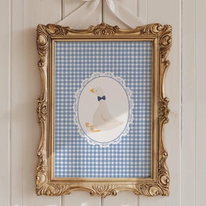 May include: A framed print with a blue and white gingham pattern. A white goose with a blue bow tie is featured in a white lace circle in the centre of the print.