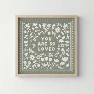 May include: Square framed print with a light wood frame. The print features the words "YOU ARE SO LOVED" in white, surrounded by a floral design on a sage green background. The floral design includes various flowers and leaves.