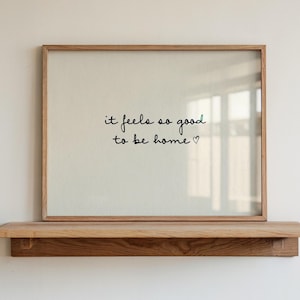 Könnte beinhalten: Ein gerahmter Druck mit dem Text "it feels so good to be home" in schwarzer Schrift. Der Druck befindet sich in einem hellbraunen Holzrahmen und ruht auf einem passenden Holzregal. Der Hintergrund ist eine schlichte weiße Wand.