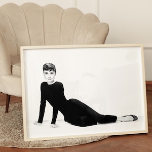 May include: Black and white photograph of a woman wearing a black dress, lying on a white background. The woman is looking directly at the camera.