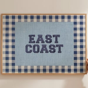 May include: A framed wall art piece with a light blue background and a navy blue gingham border. The words "EAST COAST" are embroidered in navy blue block letters. The frame is made of light-colored wood.