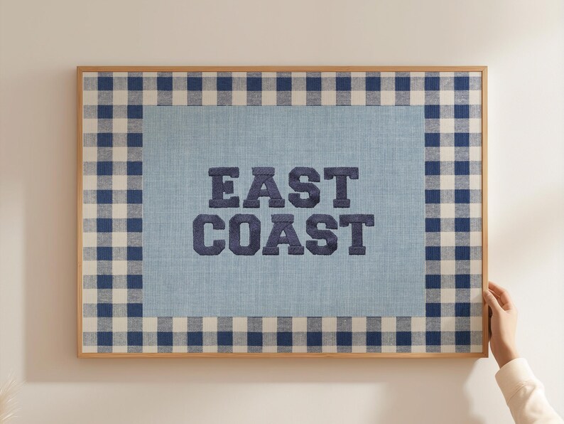 May include: A framed wall art piece with a light blue background and a navy blue gingham border. The words "EAST COAST" are embroidered in navy blue block letters. The frame is made of light-colored wood.