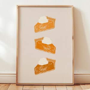 Op de afbeelding: Een ingelijste print met drie stukken pompoentaart met slagroom. De taartstukken zijn oranje met een textuur, tegen een lichtbeige achtergrond. Het frame is gemaakt van lichtgekleurd hout.