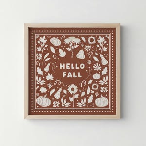 Könnte beinhalten: Quadratischer Kunstdruck mit braunem Hintergrund und weißen Illustrationen von Kürbissen, Blumen und Blättern. Die Worte "HELLO FALL" stehen in der Mitte. Der Rahmen hat eine helle Holzfarbe.