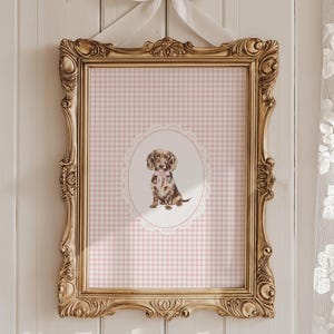 May include: An ornate gold framed print. The print features a watercolour illustration of a dachshund puppy wearing a pink bow, set against a pink and white gingham background. The frame is hanging on a white wall.