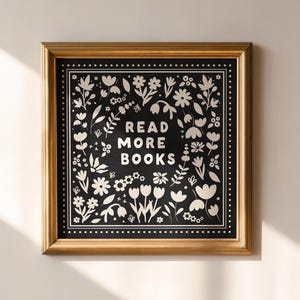 May include: A framed print with a gold-coloured frame. The artwork has a black background with white floral and leaf designs surrounding the text "READ MORE BOOKS". The square print hangs on a neutral-coloured wall.