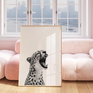 May include: A black and white photograph of a cheetah with its mouth open in a roar. The cheetah is framed in a gold frame and is hanging on a pink wall.