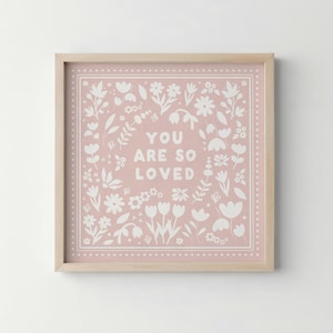May include: Square framed print with a pale pink background and white floral design. The words "YOU ARE SO LOVED" are centered. The frame is a light wood colour.