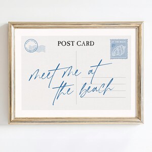 May include: Framed postcard print with the words "meet me at the beach" in blue script. The postcard design includes a postage stamp and post office markings in blue, set against a cream background.