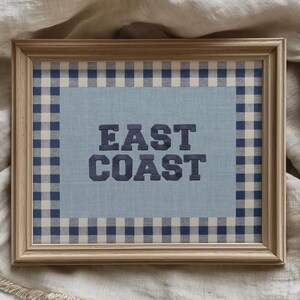 May include: Framed artwork with the words "EAST COAST" in navy blue, centered on a light blue background. The frame is a light wood color, and the artwork is surrounded by a navy and white checkered pattern.