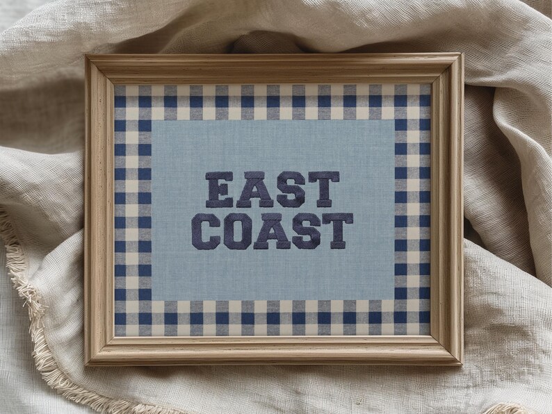 May include: Framed artwork with the words "EAST COAST" in navy blue, centered on a light blue background. The frame is a light wood color, and the artwork is surrounded by a navy and white checkered pattern.