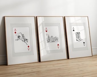 Op de afbeelding: Drie ingelijste prints van speelkaarten, elk met een ander symbool: een cowboy die een paard berijdt met een lasso, een woestijnlandschap en een paar cowboylaarzen. Elke kaart is een aas.