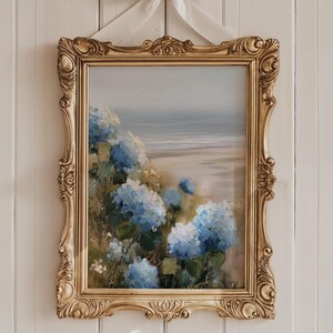 May include: A framed painting featuring a coastal landscape with blue hydrangeas in the foreground. The artwork is set in an ornate, gold-colored frame and is suspended by a white ribbon against a white wooden wall.