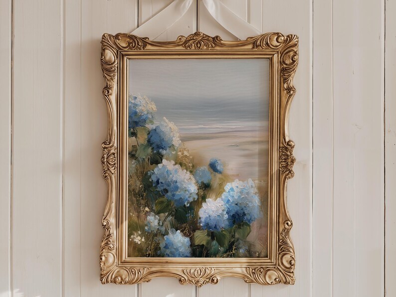 May include: A framed painting featuring a coastal landscape with blue hydrangeas in the foreground. The artwork is set in an ornate, gold-colored frame and is suspended by a white ribbon against a white wooden wall.