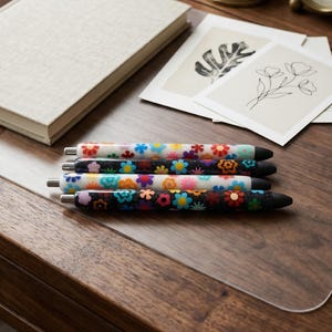 May include: Four retractable pens arranged horizontally, featuring colorful floral patterns. Two pens have a white base with multi-colored flowers, while the other two have a black, glittery base with vibrant, multi-colored flower designs.