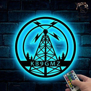 May include: A blue illuminated sign with a black silhouette of a radio tower, lightning bolts, and two pine trees. The text "KB9GMZ" is below the tower. The sign is on a brick wall with a remote control in the foreground.